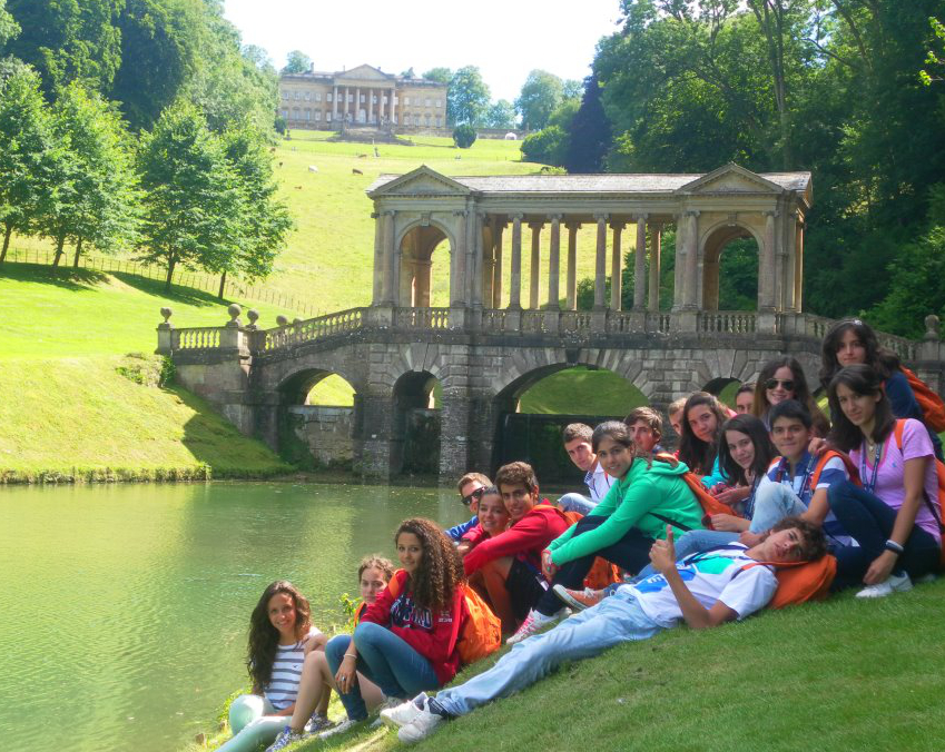 Students sitting in grass in front of a school and a bridge